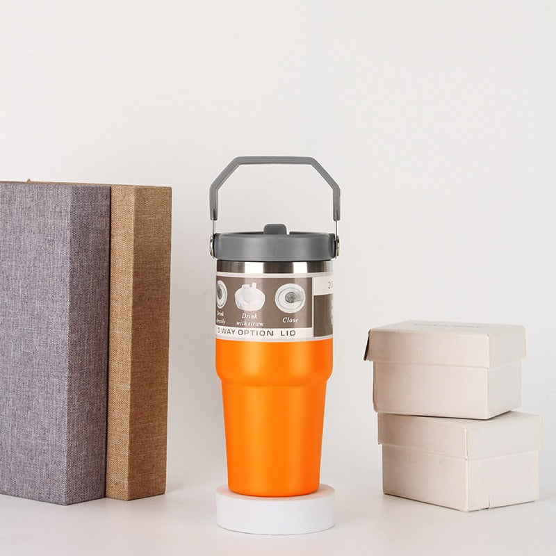 Orange insulated tumbler with gray lid and handle on a table, next to books and gift boxes, sps street style quality gifts